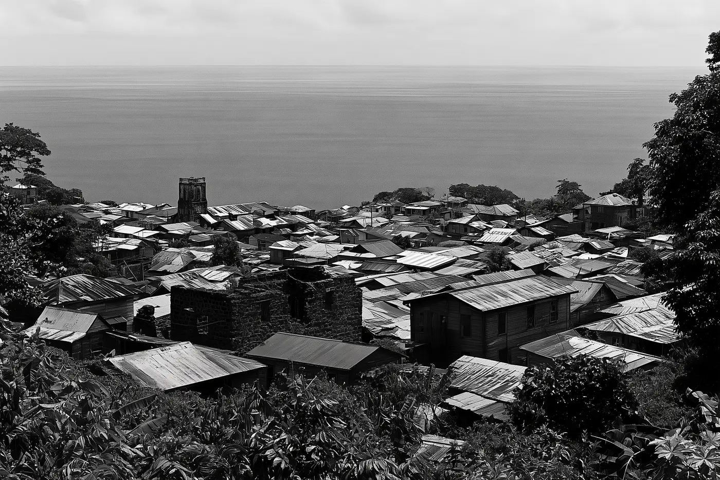 Les toits de tôle d’une petite ville côtière vus depuis la pente, des murs en ruine parmi les maisons, la mer au fond.